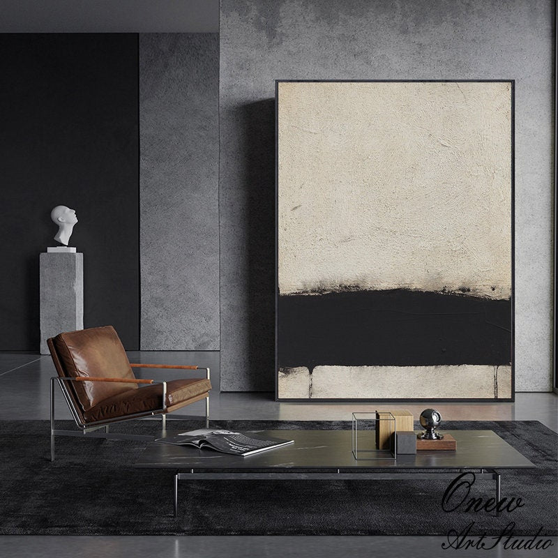 May include: Abstract art painting with a black and white color scheme. The painting features a thick black horizontal line across the middle of the canvas, with a lighter beige color above and below the line. The painting is framed in a black frame and is hanging on a wall in a modern living room.