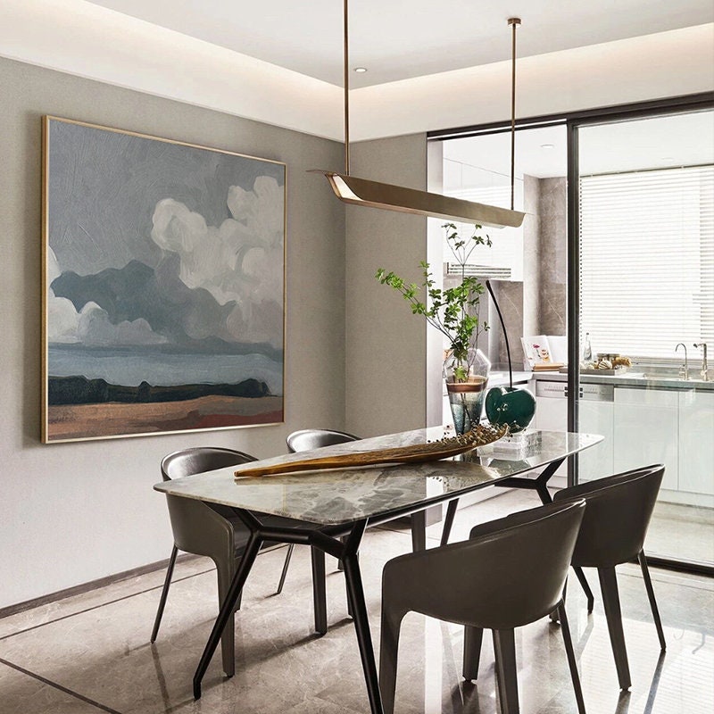 May include: An abstract landscape painting in a gold frame hangs above a dining table. The painting features a cloudy sky with shades of gray and white over a brown and blue landscape. The table has a marble top and is surrounded by four gray chairs.