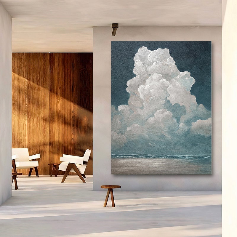 May include: Large vertical canvas art featuring a cloudscape. The painting depicts a large, fluffy white cloud against a dark blue sky. The bottom of the painting shows a light blue ocean and a sandy beach. The artwork is displayed in a modern, minimalist interior.