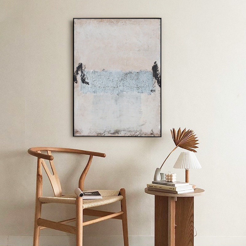 May include: A minimalist abstract painting with a light pink, white, and blue color scheme. The painting is framed in black and hangs on a white wall above a wooden chair and a side table with a lamp.