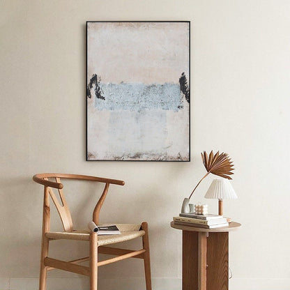 May include: A minimalist abstract painting with a light pink, white, and blue color scheme. The painting is framed in black and hangs on a white wall above a wooden chair and a side table with a lamp.