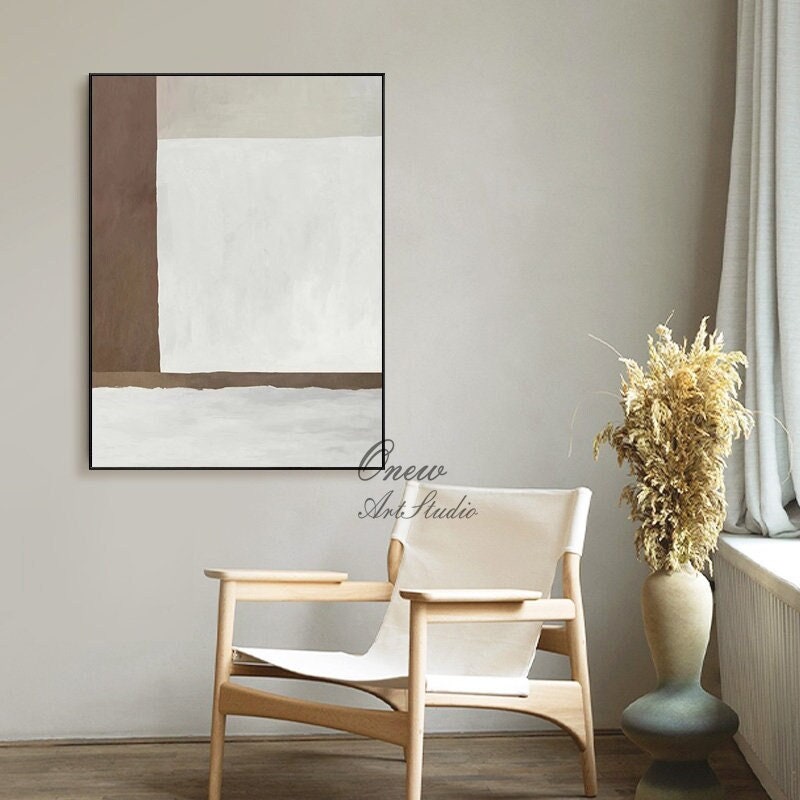 May include: Abstract painting with brown and white geometric shapes in a black frame, hanging on a wall with a white chair and a vase with dried flowers in front of it.