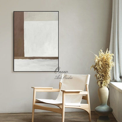 May include: Abstract painting with brown and white geometric shapes in a black frame, hanging on a wall with a white chair and a vase with dried flowers in front of it.