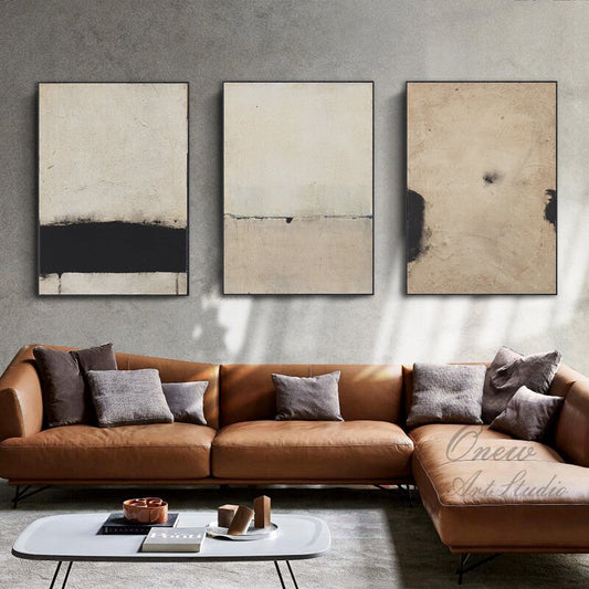 May include: Three abstract paintings in black and beige tones, hung on a gray wall above a brown leather sectional sofa. The paintings are framed in black.