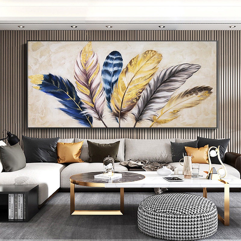 May include: A large abstract painting of gold, blue, and white feathers on a cream background. The painting is framed in a black frame and is hanging on a wall with wood paneling.