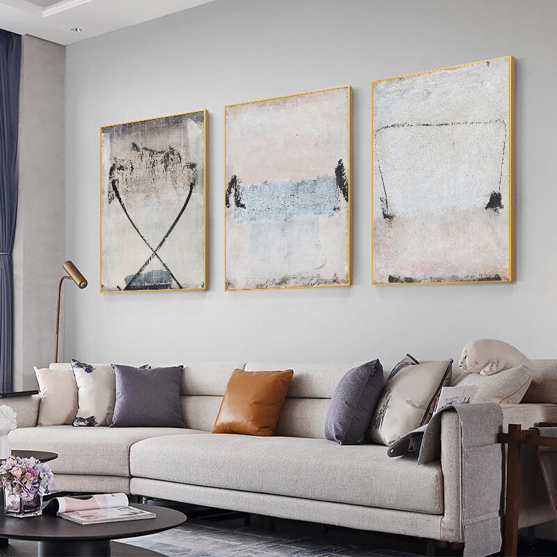 May include: Three abstract paintings with gold frames, featuring muted colors and brushstrokes. The paintings are hung on a white wall above a gray sofa with pillows.