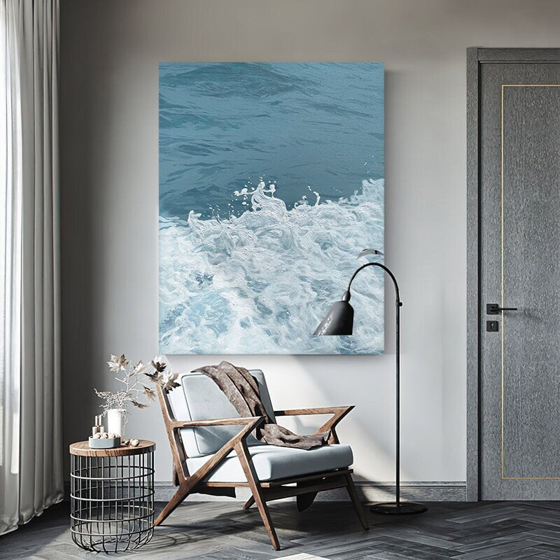 May include: A framed canvas print of a blue ocean with white foamy waves crashing. The print is hanging on a wall in a room with a gray door, a chair, and a side table.