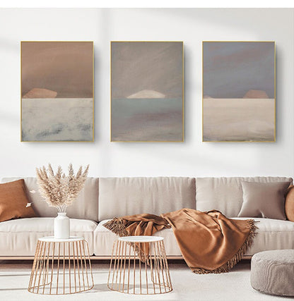 May include: Three abstract landscape paintings with muted brown, gray, and blue colors. The paintings are framed in gold and hung on a white wall above a beige sofa.
