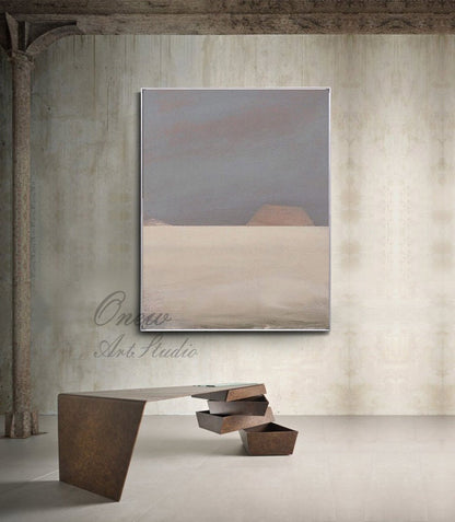 May include: Abstract painting with a gray and beige color scheme. The painting depicts a landscape with a horizon line and two hills in the distance. The painting is framed in a silver frame and is hanging on a concrete wall.
