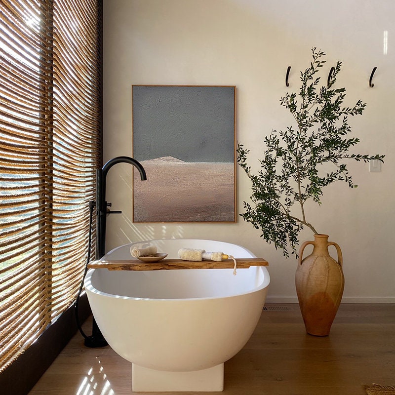 May include: A white freestanding bathtub with a wooden tray and towels. The bathtub is in a bathroom with a large window with woven blinds, a painting on the wall, and a potted olive tree.