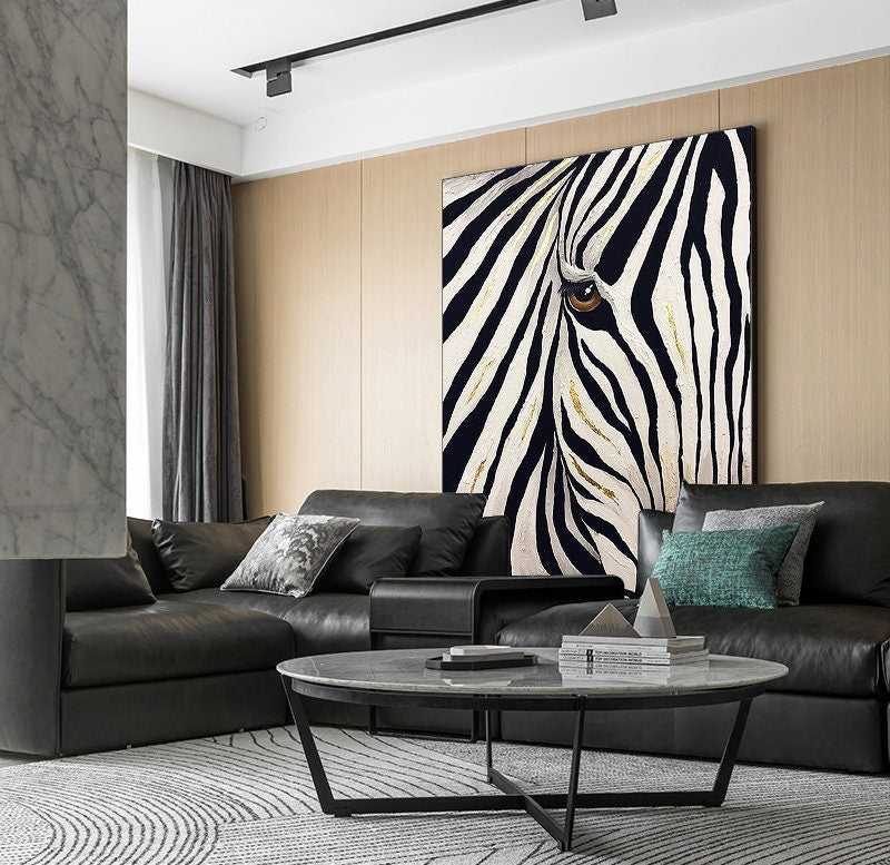 May include: A large canvas art piece featuring a close-up of a zebra's eye and black and white stripes with gold accents. The artwork hangs above a black leather sectional sofa in a modern living room setting.