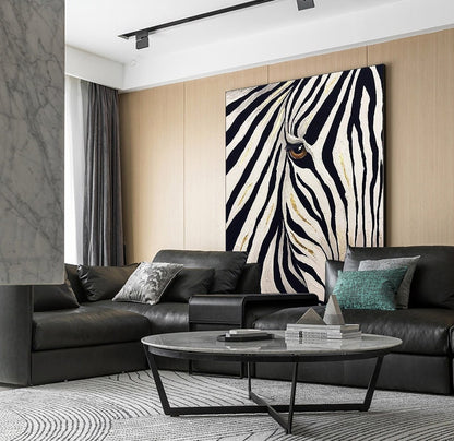 May include: A large canvas art piece featuring a close-up of a zebra's eye and black and white stripes with gold accents. The artwork hangs above a black leather sectional sofa in a modern living room setting.
