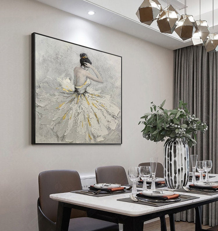 May include: A framed painting of a woman in a white dress with gold accents. The woman is standing with her head tilted down and her hands on her hair. The painting is on a white wall in a dining room setting.