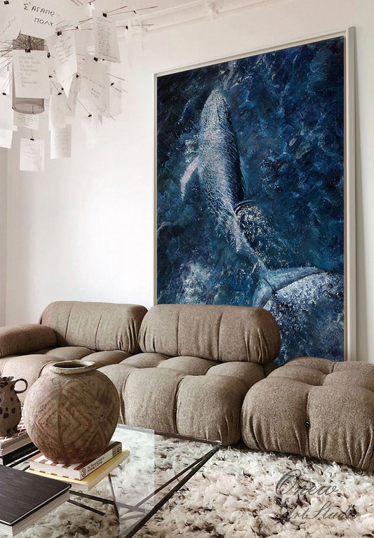 May include: A large framed painting of a whale breaching in the ocean. The whale is white and the ocean is a deep blue with white splashes of water. The painting is hanging on a white wall above a brown couch.