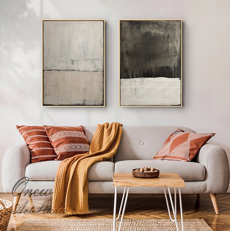 May include: Two abstract paintings in a living room setting. The paintings are both vertical and feature a horizontal line dividing the canvas into two sections. The top section of the left painting is white with gray brushstrokes, while the bottom section is a light beige. The top section of the right painting is dark gray with white brushstrokes, while the bottom section is white. The paintings are hanging on a white wall above a gray sofa with orange and white throw pillows and a brown blanket. A wooden