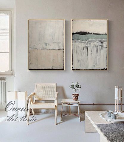 May include: Two abstract paintings in neutral colors with a light brown, gray, and green color scheme. The paintings are framed in a light wood frame and are hanging on a light gray wall.