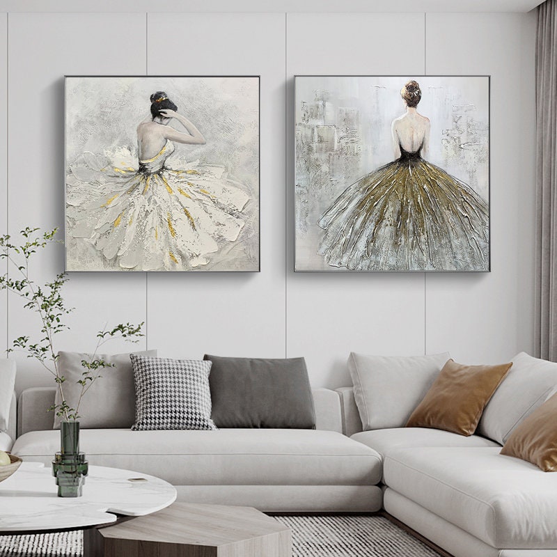 May include: Two square canvas paintings of a ballerina in a white and gold dress. The ballerina is facing away from the viewer in both paintings. The paintings are hung on a white wall above a white couch.