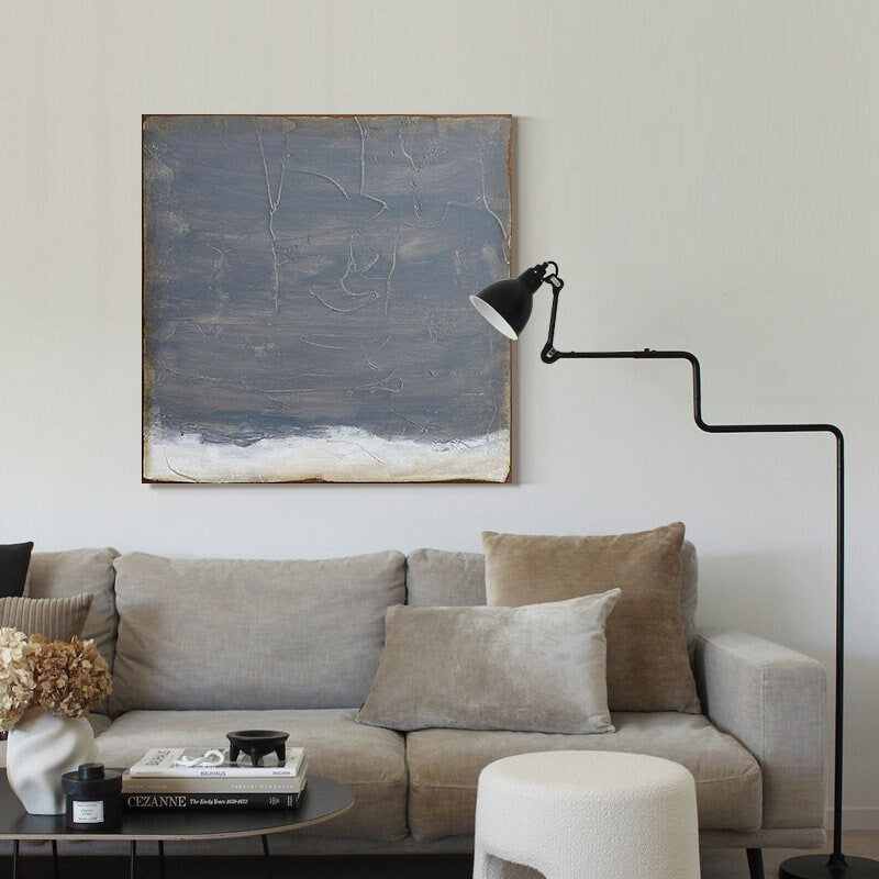 May include: A gray and white abstract painting hangs above a gray sofa with brown and beige pillows. A black floor lamp with a white shade is in the corner of the room.