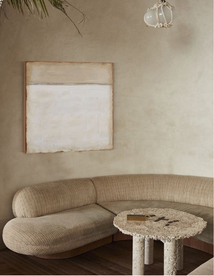 May include: A curved beige sofa with a textured fabric sits in a room with a beige wall and a small, round, textured stone table with four legs. A white light fixture hangs from the ceiling.