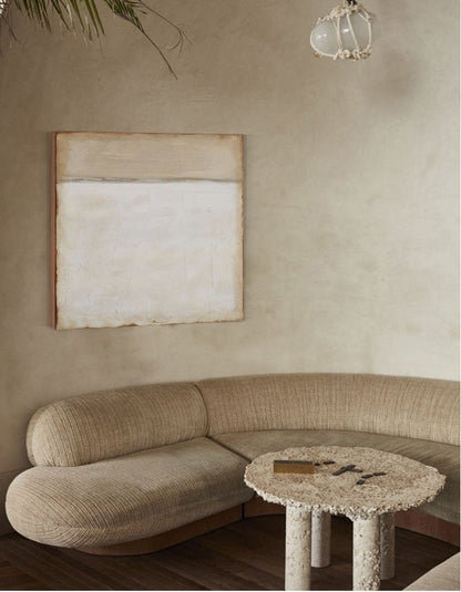 May include: A curved beige sofa with a textured fabric sits in a room with a beige wall and a small, round, textured stone table with four legs. A white light fixture hangs from the ceiling.