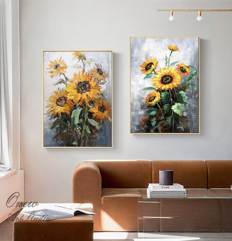 May include: Two framed paintings of sunflowers in a contemporary living room setting. The paintings are in a muted color palette with shades of yellow, green, and gray. The paintings are hung on a white wall above a brown sofa.