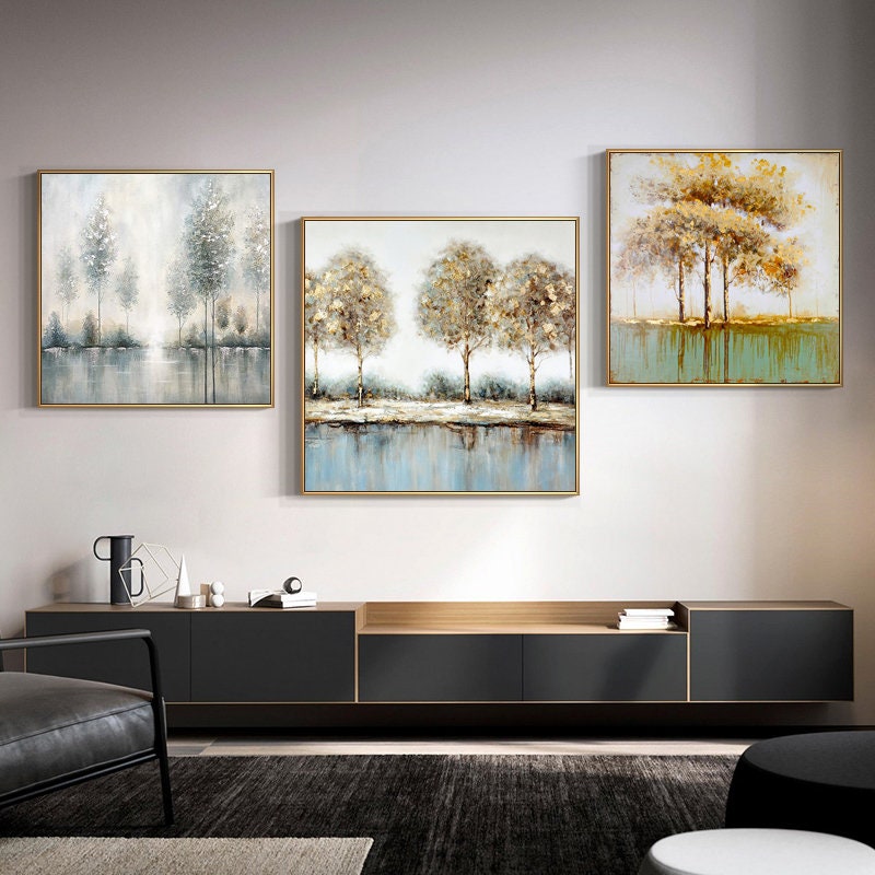 May include: Three framed abstract landscape paintings with a muted color palette of gold, brown, and blue. The paintings depict trees and water, with a focus on the reflection of the trees in the water. The paintings are hung on a white wall above a black entertainment center.