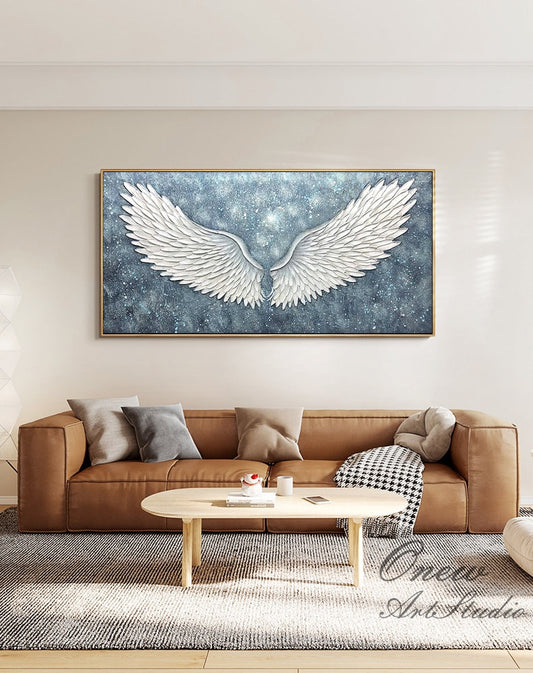 May include: A large abstract painting of white angel wings on a blue background with a glittery texture. The painting is framed in gold and is hanging on a white wall above a brown leather sofa.