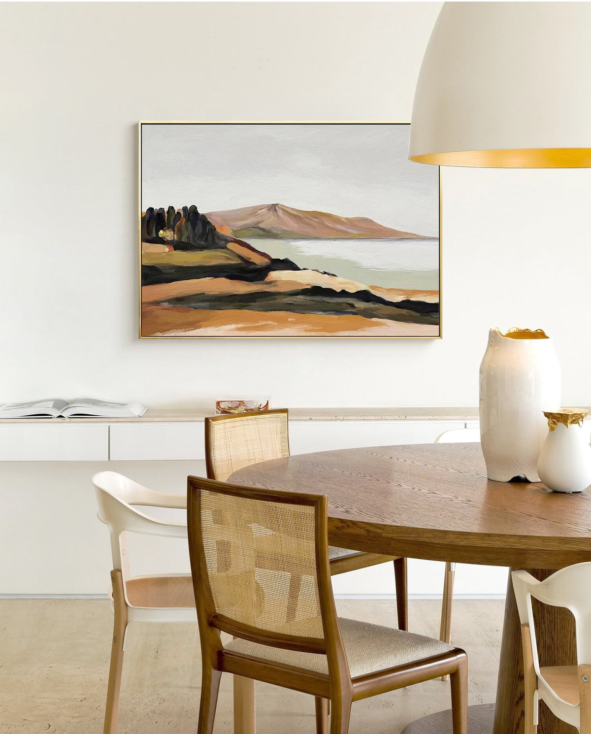 May include: A framed abstract landscape painting with a muted color palette of brown, green, and gray. The painting depicts a mountain range, a body of water, and a shoreline with trees. The painting is hanging on a white wall above a wooden table with chairs.
