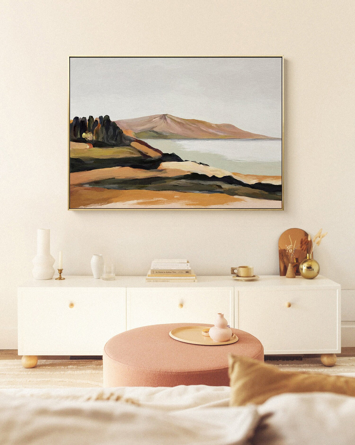 May include: A framed abstract landscape painting with a mountain range, a body of water, and a small cluster of trees in the foreground. The painting is in shades of brown, green, and blue, and is hung on a white wall above a white cabinet.