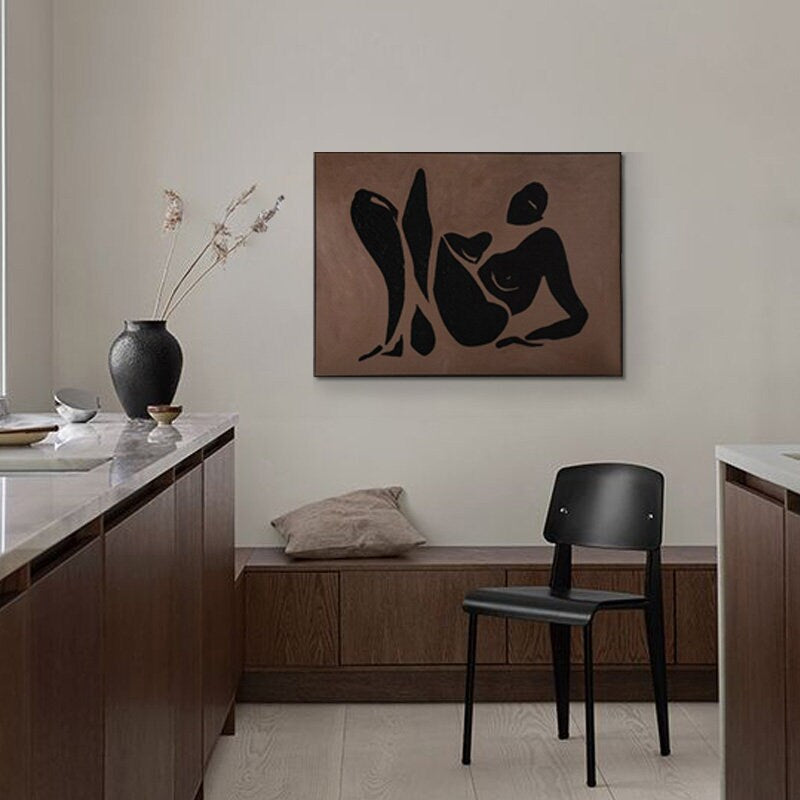 May include: A black and white abstract art print of a person in a reclining pose on a brown background. The print is hanging on a wall in a modern kitchen with wooden cabinets and a black chair.