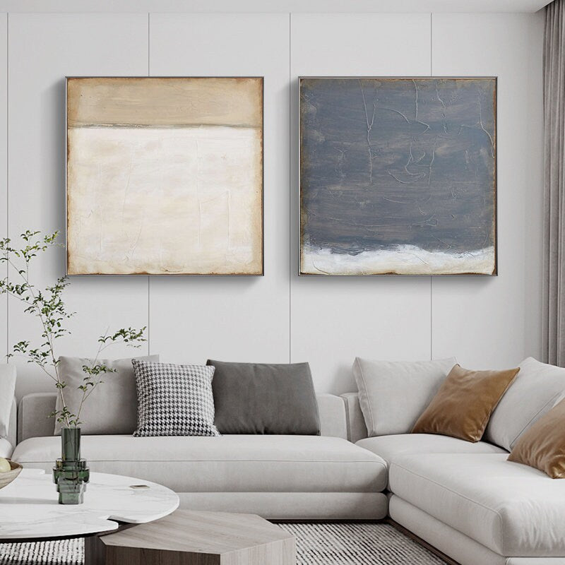 May include: Two abstract paintings in a living room setting. The painting on the left is a white and beige abstract with a horizontal line. The painting on the right is a dark blue and white abstract with a horizontal line. The paintings are hanging on a white wall above a white sectional sofa with pillows.