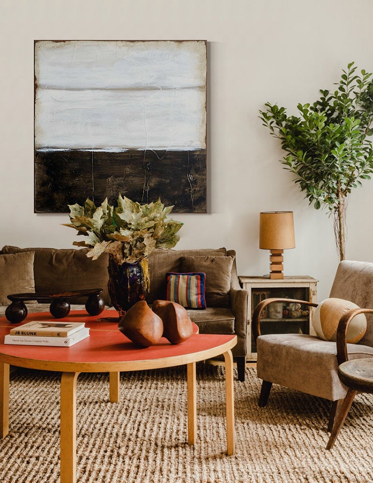 May include: A living room interior with a brown sofa, a wooden coffee table, a brown armchair, a potted plant, and a painting with a white and black abstract design.