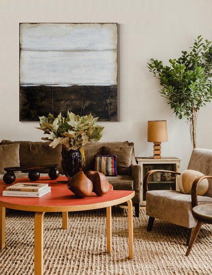 May include: A living room interior with a brown sofa, a wooden coffee table, a brown armchair, a potted plant, and a painting with a white and black abstract design.
