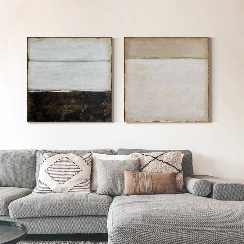 May include: Two abstract paintings with horizontal lines in shades of white, brown, and beige. The paintings are framed and hung on a white wall above a gray sectional sofa with pillows.