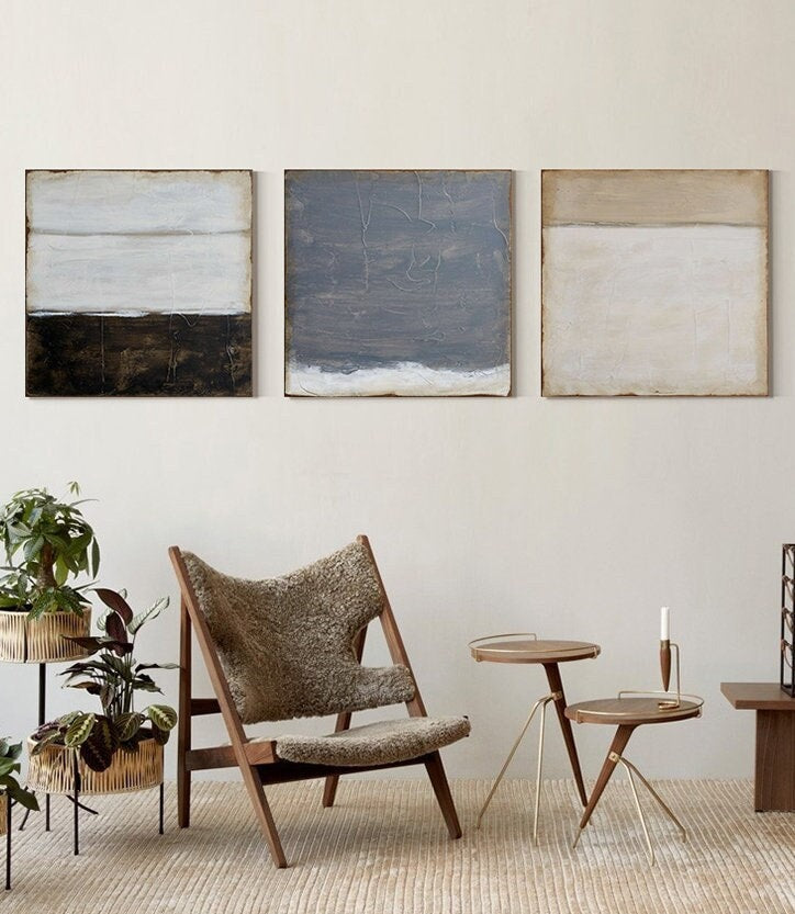 May include: Three abstract paintings with horizontal lines in shades of white, brown, gray, and beige. The paintings are hanging on a white wall above a brown chair and two small gold metal side tables.