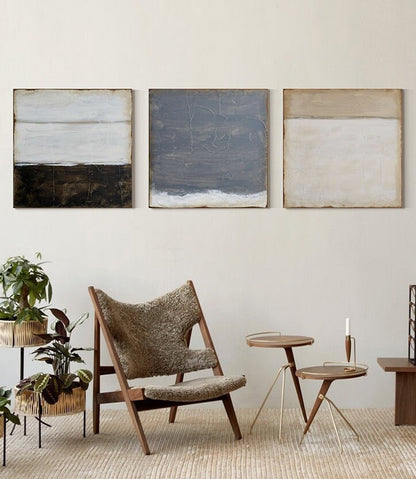 May include: Three abstract paintings with horizontal lines in shades of white, brown, gray, and beige. The paintings are hanging on a white wall above a brown chair and two small gold metal side tables.