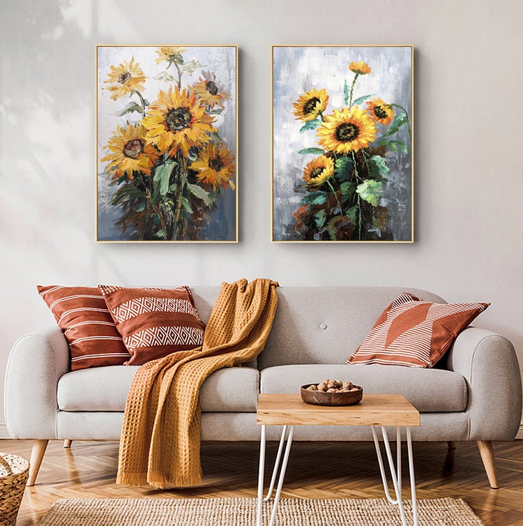 May include: Two framed paintings of sunflowers with a gray background. The paintings are hanging on a white wall above a gray couch with throw pillows and a blanket.