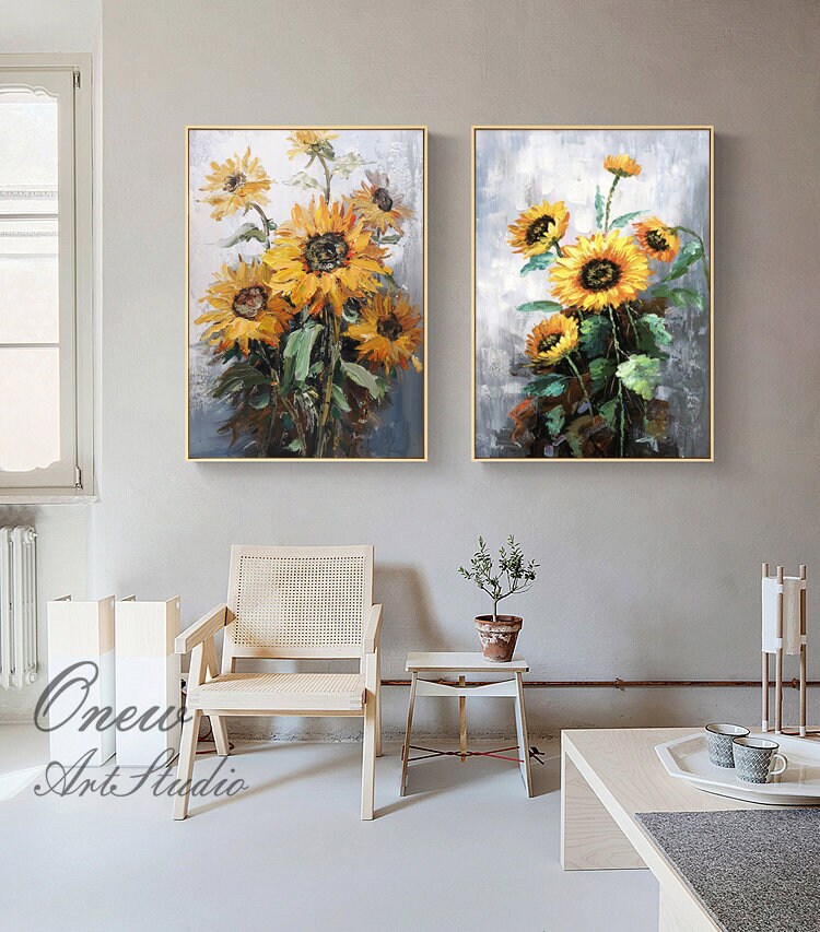 May include: Two framed paintings of sunflowers in a living room setting. The paintings are on a gray wall with a light wood chair and table in front of them. The sunflowers are painted in shades of yellow, orange, and green.