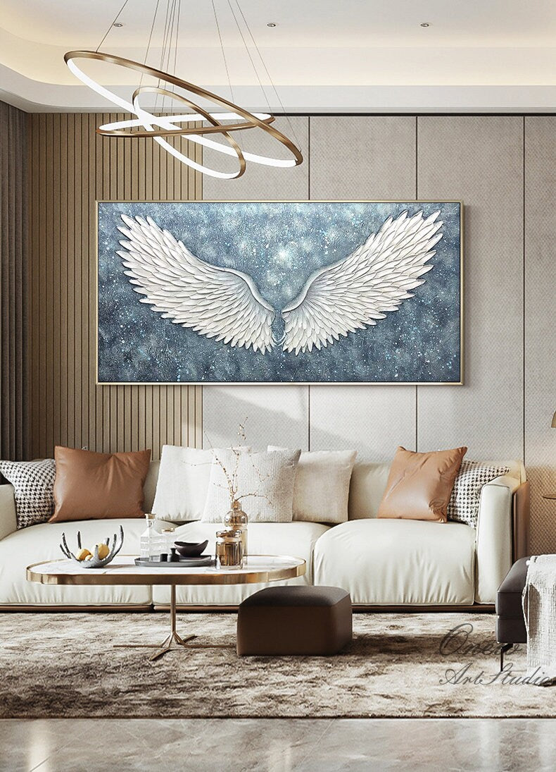 May include: A large white canvas painting of a pair of angel wings against a blue and silver background. The wings are textured and appear to be 3D. The painting is hanging on a wall in a living room setting.