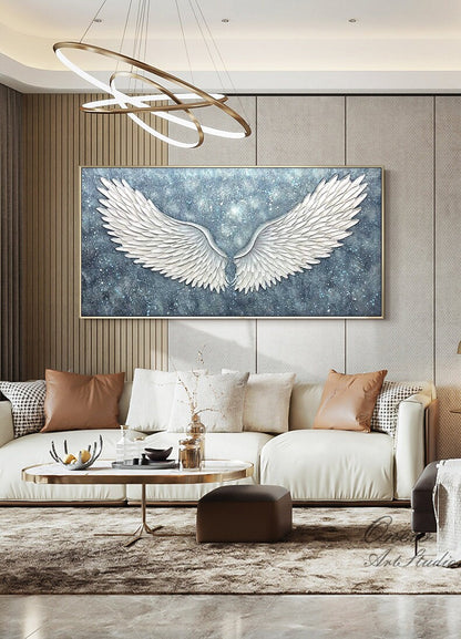 May include: A large white canvas painting of a pair of angel wings against a blue and silver background. The wings are textured and appear to be 3D. The painting is hanging on a wall in a living room setting.