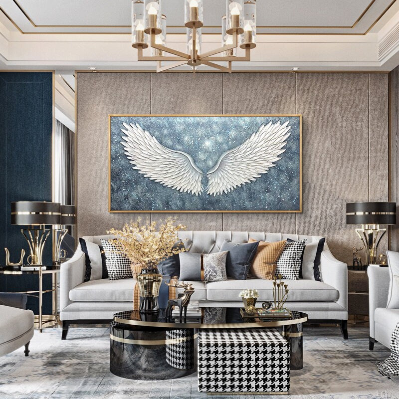 May include: A white, textured, angel wing canvas painting with a blue and white starry background. The painting is hanging on a wall in a living room with a white sofa, a black and white patterned ottoman, and a black coffee table.