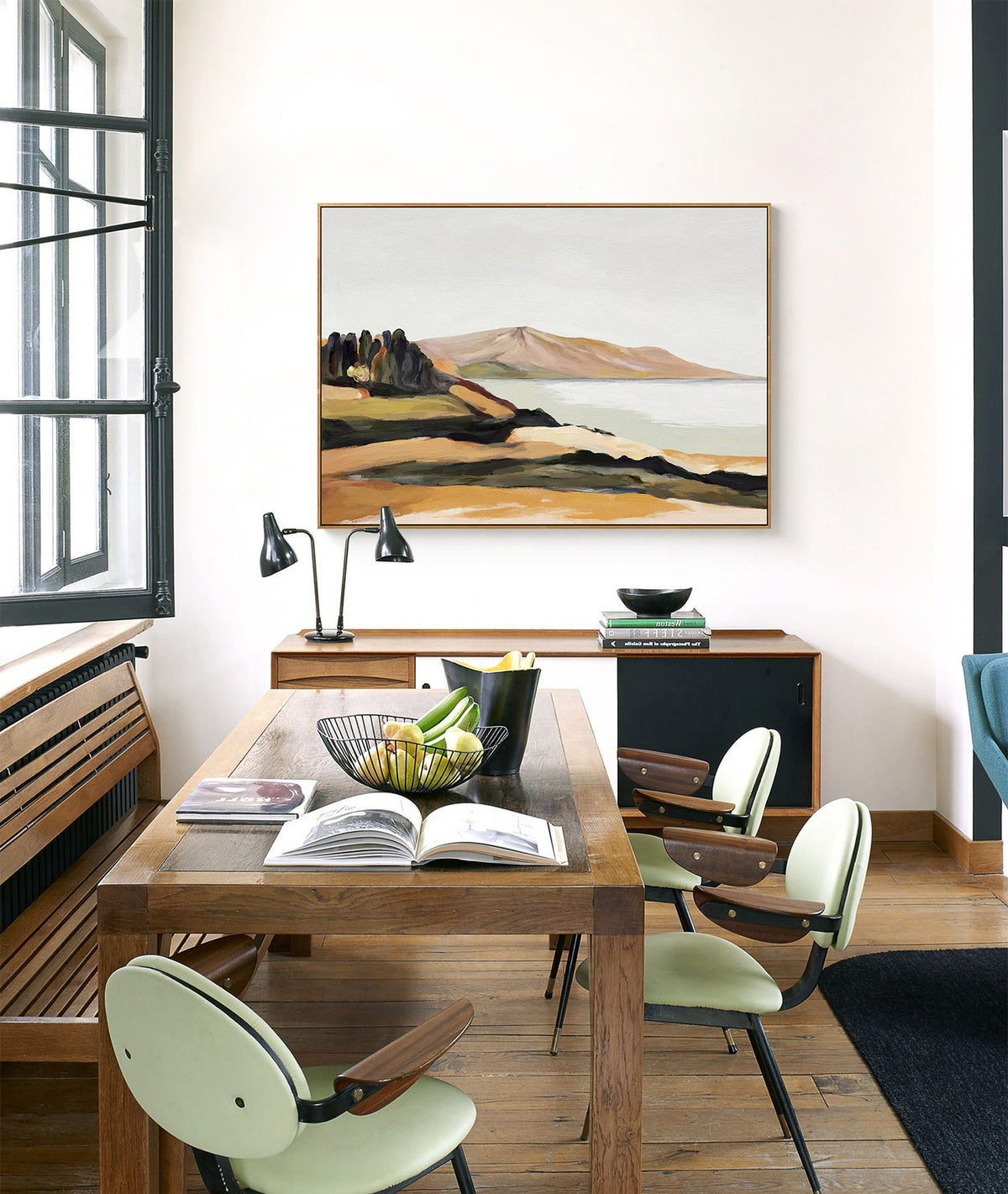 May include: A dining room with a wooden table and chairs. The table has a bowl of fruit and a book on it. There is a painting of a landscape on the wall behind the table.