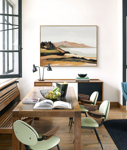 May include: A dining room with a wooden table and chairs. The table has a bowl of fruit and a book on it. There is a painting of a landscape on the wall behind the table.