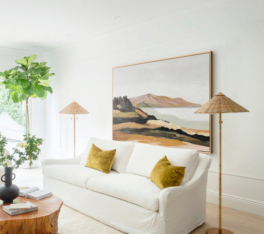 May include: A white sofa with two yellow throw pillows in a living room with a large abstract landscape painting on the wall and a woven lampshade floor lamp.