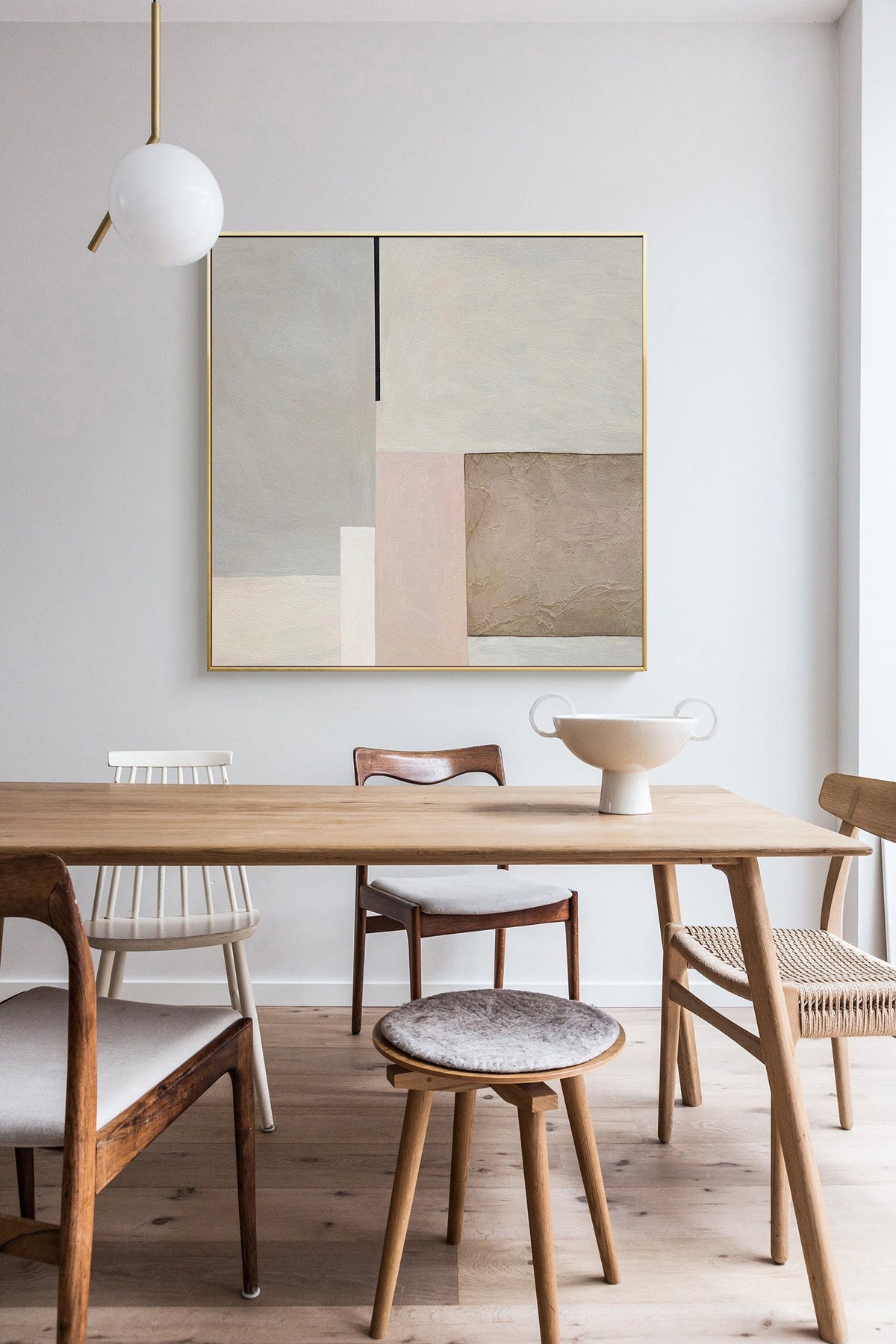 May include: A modern abstract painting with geometric shapes in shades of beige, brown, and pink hangs above a wooden dining table with chairs. The painting is framed in gold.