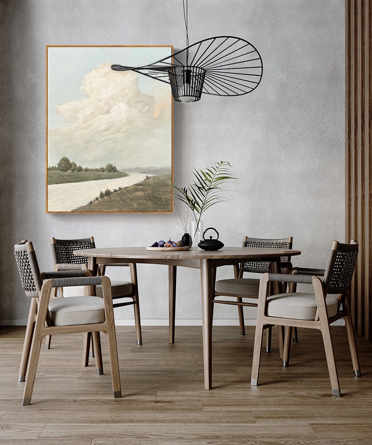 May include: A dining room with a wooden table and chairs. The table is round and has a light wood finish. The chairs are also made of wood and have a woven seat. There is a painting of a landscape with a river and clouds hanging on the wall. A black metal light fixture hangs above the table.