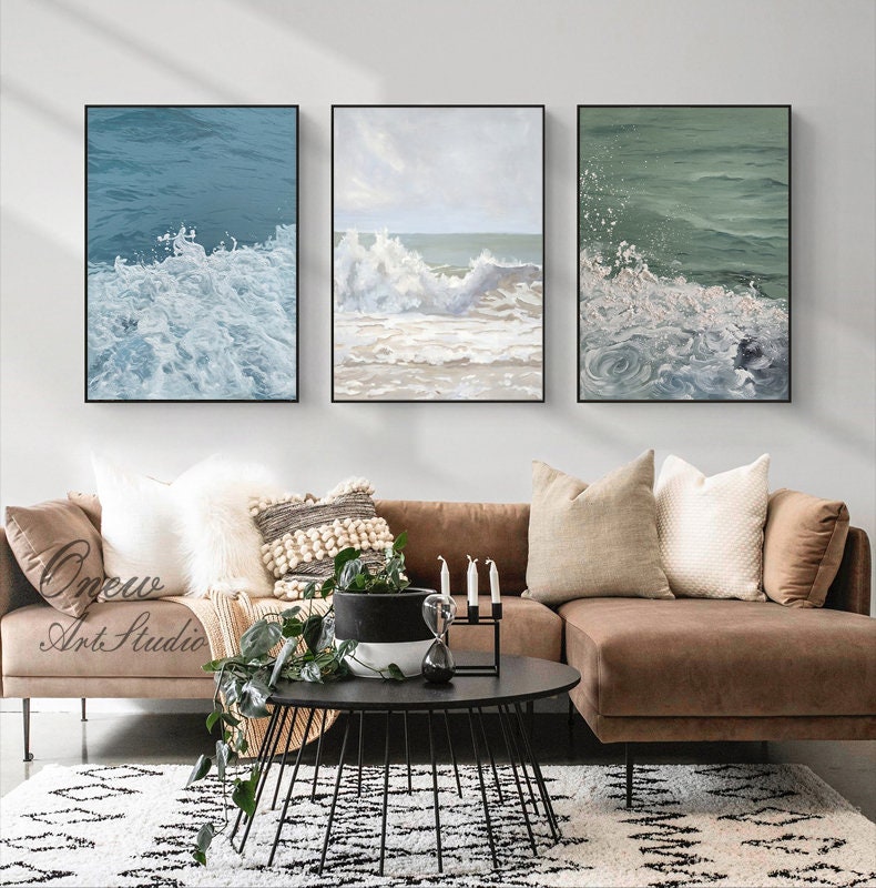 May include: Three framed canvas paintings depicting ocean waves crashing on the shore. The paintings are in a living room setting with a brown sectional sofa, a coffee table, and a potted plant.