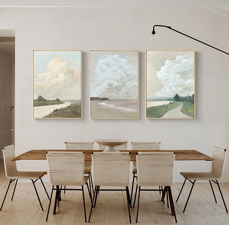 May include: Three framed landscape paintings with a minimalist style. Each painting features a large, fluffy cloud in a light blue sky over a green field and a body of water. The paintings are hung on a white wall above a wooden dining table with six chairs.