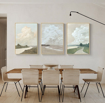 May include: Three framed landscape paintings with a minimalist style. Each painting features a large, fluffy cloud in a light blue sky over a green field and a body of water. The paintings are hung on a white wall above a wooden dining table with six chairs.