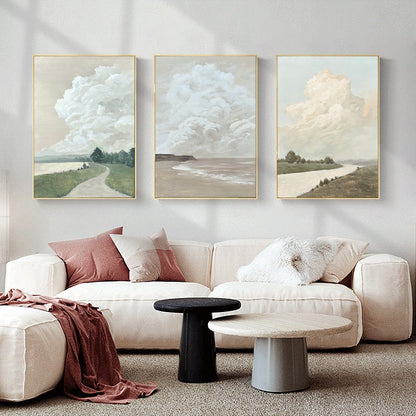 May include: Three framed canvas prints with a minimalist landscape theme. Each print features a large, fluffy cloud in a pale blue, white, or cream color against a light beige background. The bottom of each print shows a different landscape scene: a path through a grassy field, a beach with waves, and a river winding through a field.